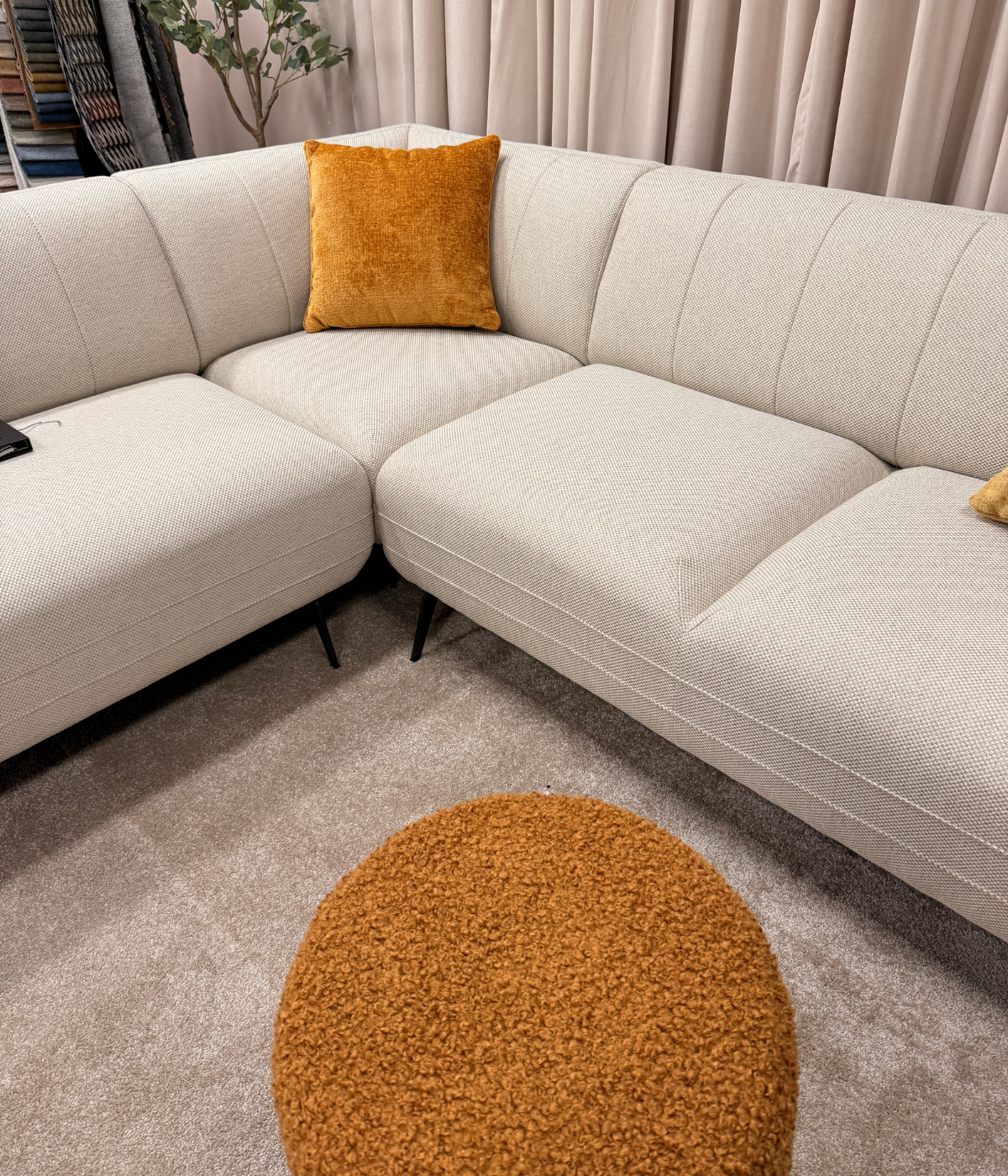 Narożnik Rea z pomarańczową pufą Leticia na tle beżowego dywanu i zasłony