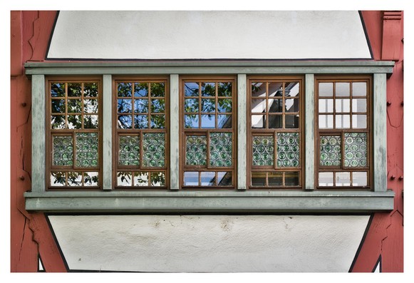 The picture shows a large, wide window with several small panes of glass embedded in a white wall with red wooden beams. The window has a gray frame and consists of five vertical sections. The top three quarters are clear glass panes showing daylight and reflections of trees and sky. Below are decorative glass panels that have a pattern of circular green and white ornaments arranged like small tiles. The lowest section is again made of clear glass panes. The window looks old and slightly weathered, with visible signs of wear on the frame.