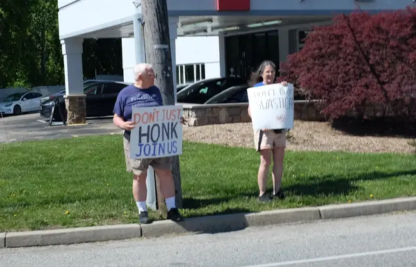 A man holds a poster saying "Don't just honk, join us." His wife, to the right, holds a poster saying "Don't buy swasticars."