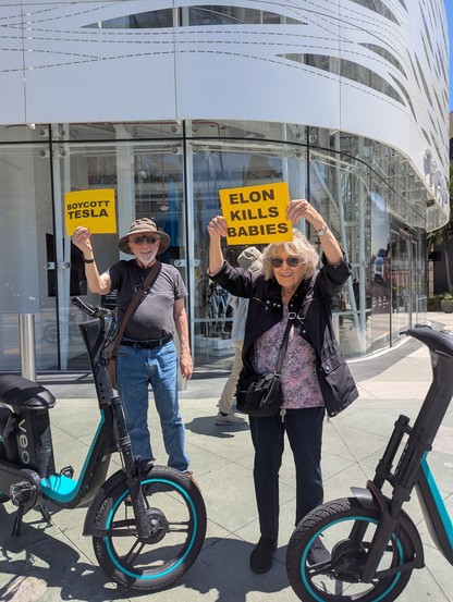 A grey haired man and woman standing on a street corner holding up protest signs reading "Boycott Tesla" and "Elon Kills Babies"