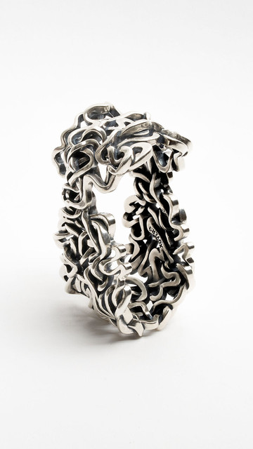 Xover0's solid sterling silver napkin ring stands upright on a white background. This functional sculpture was created by interweaving seven ancient courses of the Mississippi River. The resulting forms are ornate and organic, capturing fluid dynamics in solid form. With bold dimensionality and a commanding presence, their liquid silver surfaces scatter light and evoke a sense of motion.
