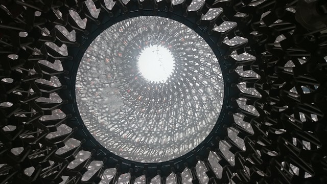 Looking directly up into a large, intricate architectural structure from the Milan World Expo in 2015, featuring a dense pattern of interconnected elements that frame a bright view of the sky above.