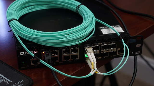 A QNAP 10GbE switch with two SFP+ modules plugged in and a OM3 LC/LC fiber optic cable connecting the two together.  The switch is very much not happy in the photo.