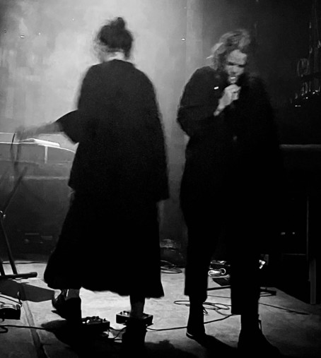 Monasunne performing live on a dark stage. One performer stays with the back to the camera, in front of a synthesizer. The other performer looks down, singing into a microphone. There are effect pedals on the floor.
