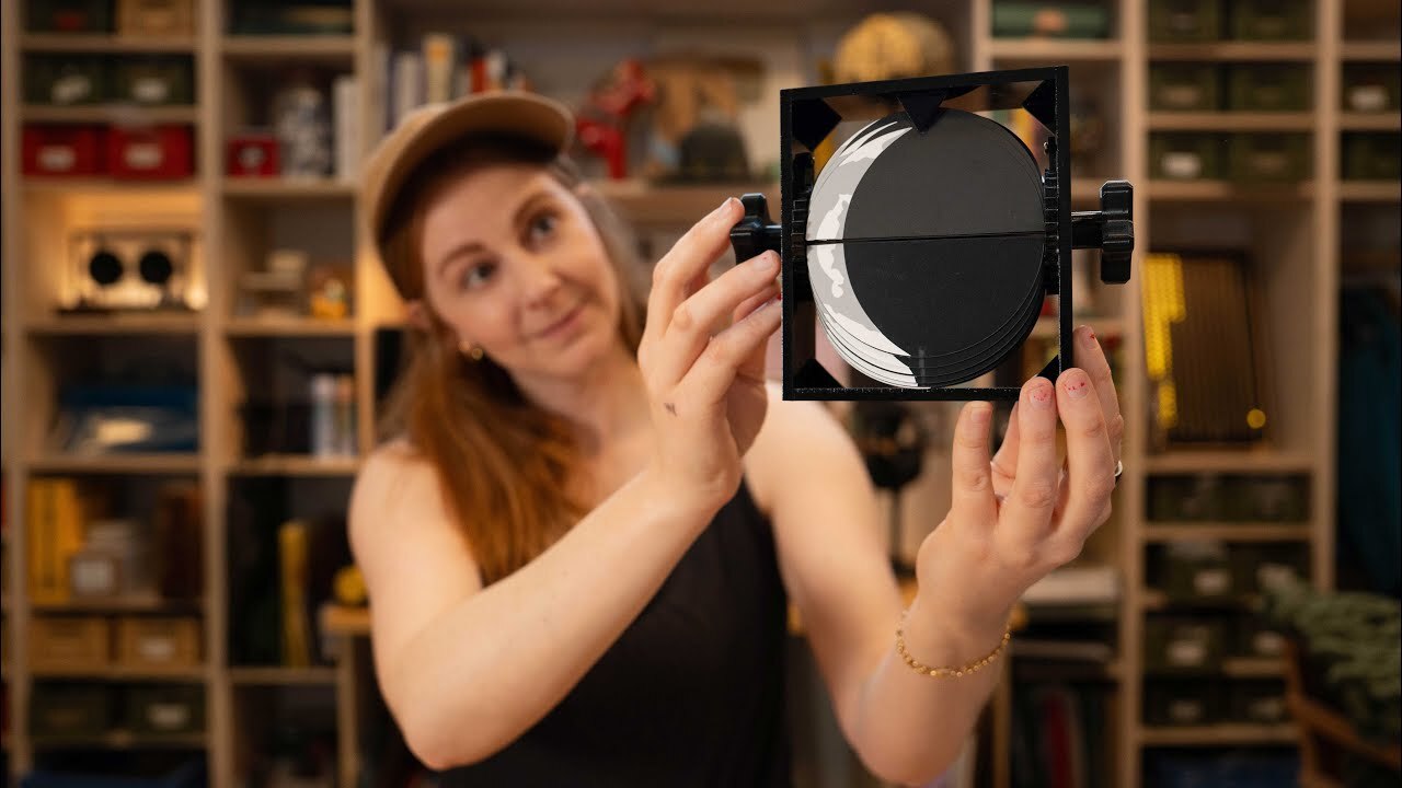 A woman in a ball cap and black tank top holds a black and white image of the Moon printed on black acrylic. It can fold and is mounted on a black plastic mechanism with turning knobs affixed to the ends. There are out-of-focus shelves in the background with various items and books on them. A woman in a ball cap and black tank top holds a black and white image of the Moon printed on black acrylic. It can fold and is mounted on a black plastic mechanism with turning knobs affixed to the ends. There are out-of-focus shelves in the background with various items and books on them.