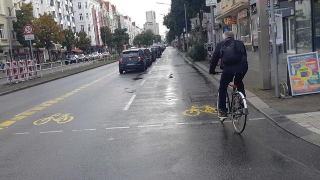 Ein Radfahrer fährt auf dem Pop-up-Radweg in der Kantstraße. Links davon die kfz-Stellplätze. Es hat geregnet, der Asphalt ist nass.