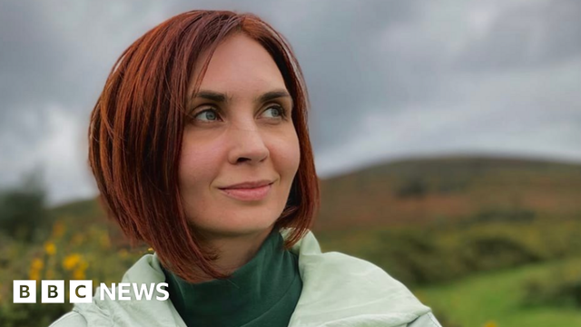 A lady stood outdoors with hills and greenery in the background. She has short, reddish-brown hair and is wearing a light green jacket over a dark green top. They are holding the collar of the jacket with both hands.