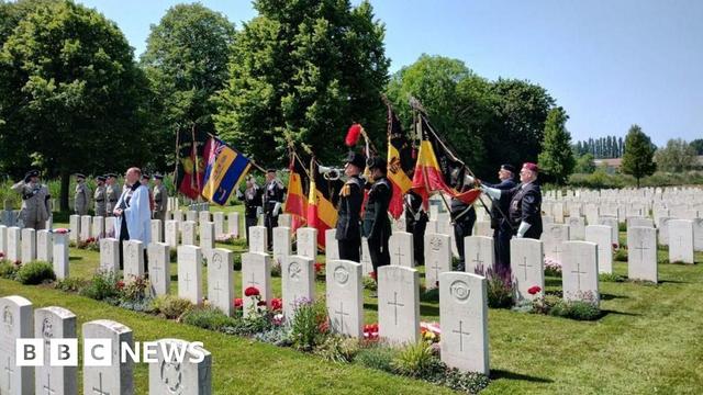 A padre wearing a white cloak and men in military uniform holding up flags, standing in a cemetery with neat rows of white graves. It's a sunny day and the sky is blue.