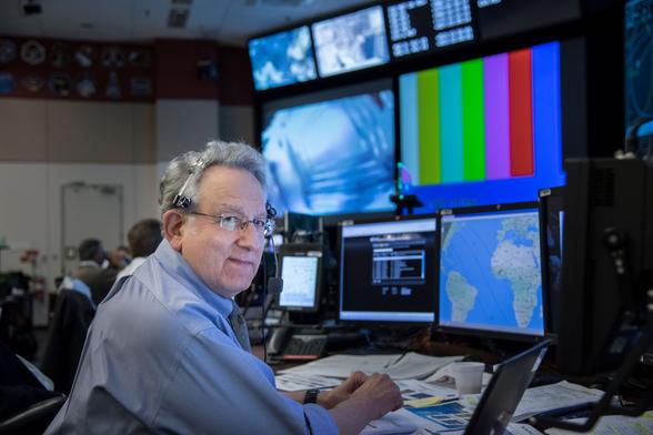 A man on console sits in front of several monitors and a desk covered in paperwork.