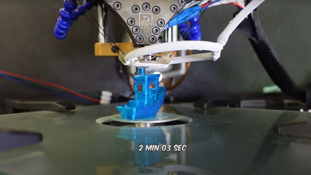 A blue 3DBenchy is visible on a small circular plate extending up through a cutout in a flat, reflective surface. Above the Benchy is a roughly triangular metal 3D printer extruder, with a frost-covered ring around the nozzle. A label below the Benchy reads “2 MIN 03 SEC.”