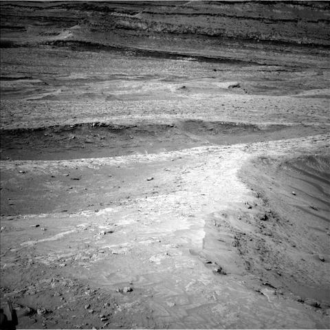 A grayscale photo from the Martian surface shows rocky, uneven, light-gray terrain marked by numerous ridges, running at various angles to each other, creating lines atop the sandy, gravelly soil.
