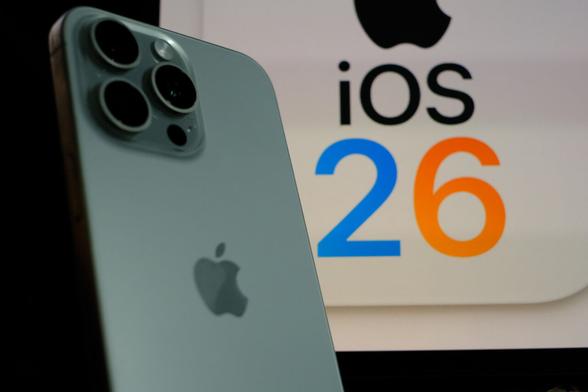 Tylna część srebrnego iPhone’a z widocznym modułem trzech aparatów i logo Apple na rozmytym tle wyświetlającym napis iOS 26 (2 niebieska, 6 pomarańczowa).