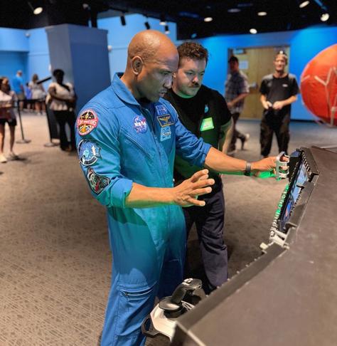 An astronaut in a blue NASA flight suit leans forward to interact with an electronic screen on exhibit about the Orion spacecraft. A man beside him looks on. People look on from a distance.