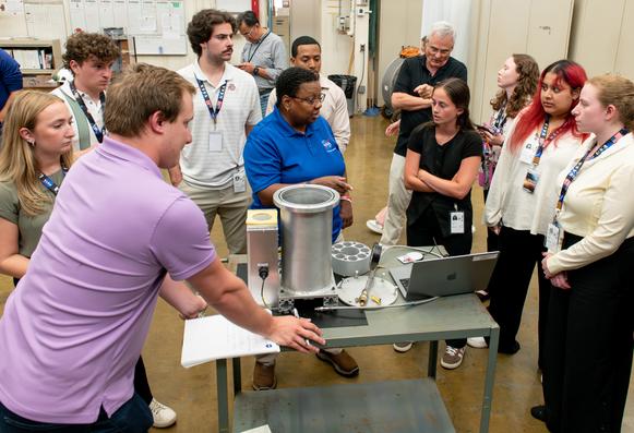 Several students surround a table containing steel components and wires as they listen to a Glenn engineer explain the technology.