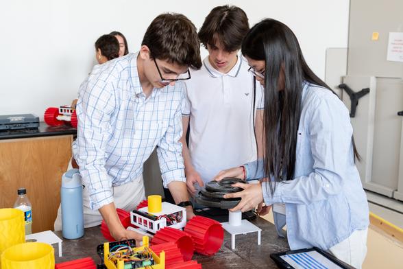Three students stand next to a table and carefully prepare to place weights on top of a plastic rover with large red wheels. Additional plastic rover parts and wires sit on the table. Two students are in the background discussing their rover, which sits on a counter.