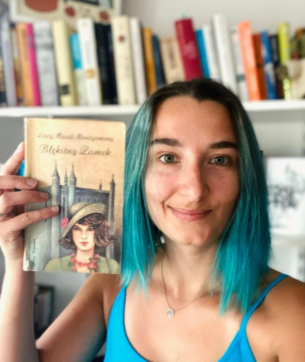 Kasika pozująca z książką Lucy Maud Montgomery pt. Błękitny zamek. Zdjęcie portretowe, zrobione na tle półek z książkami. Wydanie książki stare, zżółknięte, podniszczone. Kasika ma niebieskie włosy i jest uśmiechnięta (widać, że opinia o książce będzie pozytywna)