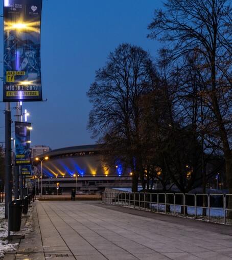 Widok na ulicę w kierunku Spodka, po lewej stronie kadru bannery przedstawiające mistrzów poprzednich edycji wiszące na słupach oświetleniowych wzdłuż chodnika, po prawej kilka drzew