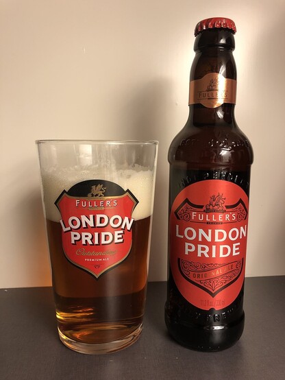 zdjęcie butelki i szklanki piwa Fuller's London Pride, z czerwonymi etykietami w stylu tarczy herbowej, szklanka pełna ciemno-złotego piwa z grubą pianą, stoją na tle białej ściany