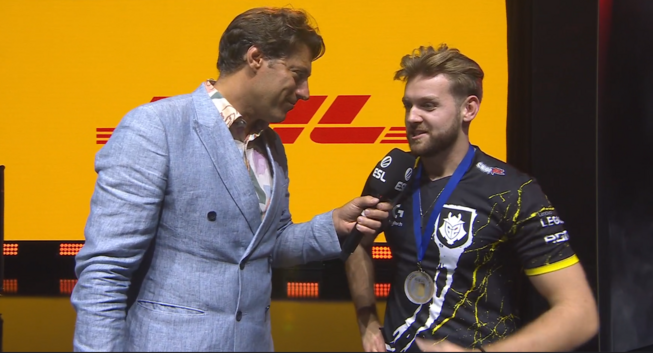 zawodnik G2 Esports, NiKo w wywiadzie pomeczowym z hostem sceny IEM Cologne, NiKo trzyma w rękach nagrodę za MVP turnieju, w tle telebim z reklamą DHL na pomarańczowym tle