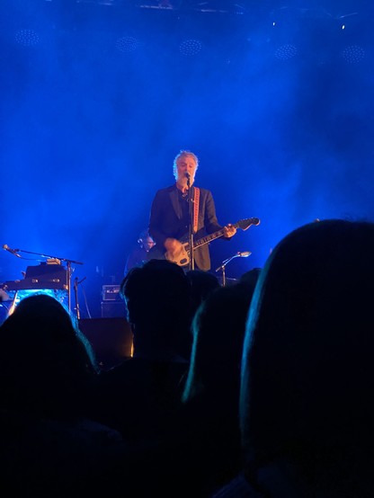 Glen Hansard na scenie z gitarą, tło niebieskie (światło reflektorów), z przodu na dole trochę głów ludzi (zdjęcie robione z tłumu pod sceną)