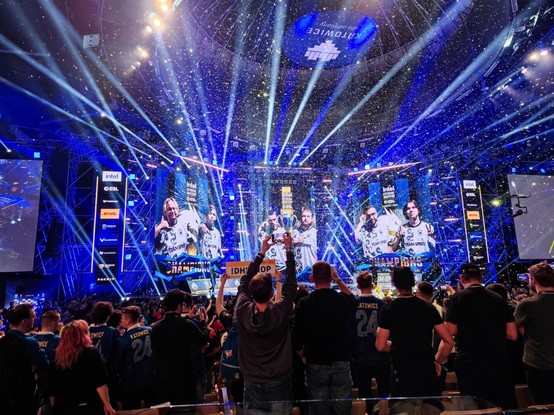 scena katowickiego Spodka rozświetlona na niebiesko i biało, na ekranach widoczni zawodnicy Team Spirit w białych koszulkach meczowych z podpisem Champions, na pierwszym planie stojący kibice, z sufitu sypie się złote konfetti