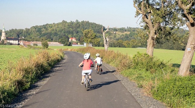 Dwoje dzieci jeździ na rowerach wiejską ścieżką, otoczoną zielonymi polami i odległymi wzgórzami. Mała wioska z budynkami i wieżą kościelną jest widoczna w tle. Scena rozgrywa się w słoneczny dzień.