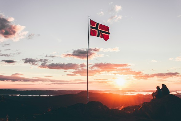 Krajobraz górski z zachodzącym słońcem i flagą Norwegii. Źródło: https://unsplash.com/photos/norway-flag-standing-on-cliff-HO6aBrYi3kE