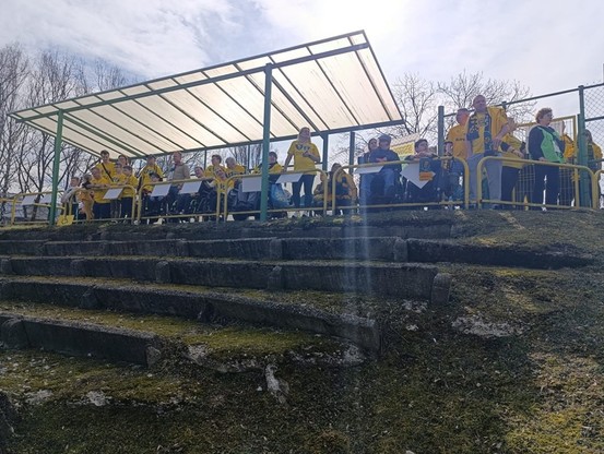 Zdjęcie przedstawia grupę niepełnosprawnych kibiców GKS Katowice, w większości ubranych w żółte koszulki klubowe, znajdujących się na trybunie dla osób z niepełnosprawnościami zaadaptowanej ze starych, wykonanych z kamienia trybun stadionowych. Trybuna jest mała, zadaszona, wyposażona w barierki i oznaczenia miejsc dla wózków inwalidzkich. W tle widoczne są bezlistne drzewa i słoneczne niebo.