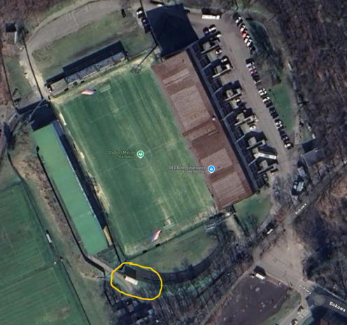 Zdjęcie lotnicze przedstawiające stary Stadion Miejski w Katowicach z widoczną płytą główną boiska, trybunami, parkingiem oraz sąsiadującymi boiskami treningowymi i drogami. Zaznaczona na żółto jest malutka trybunka dla osób niepełnosprawnych, ulokowana w rogu, trochę za linią końcową.