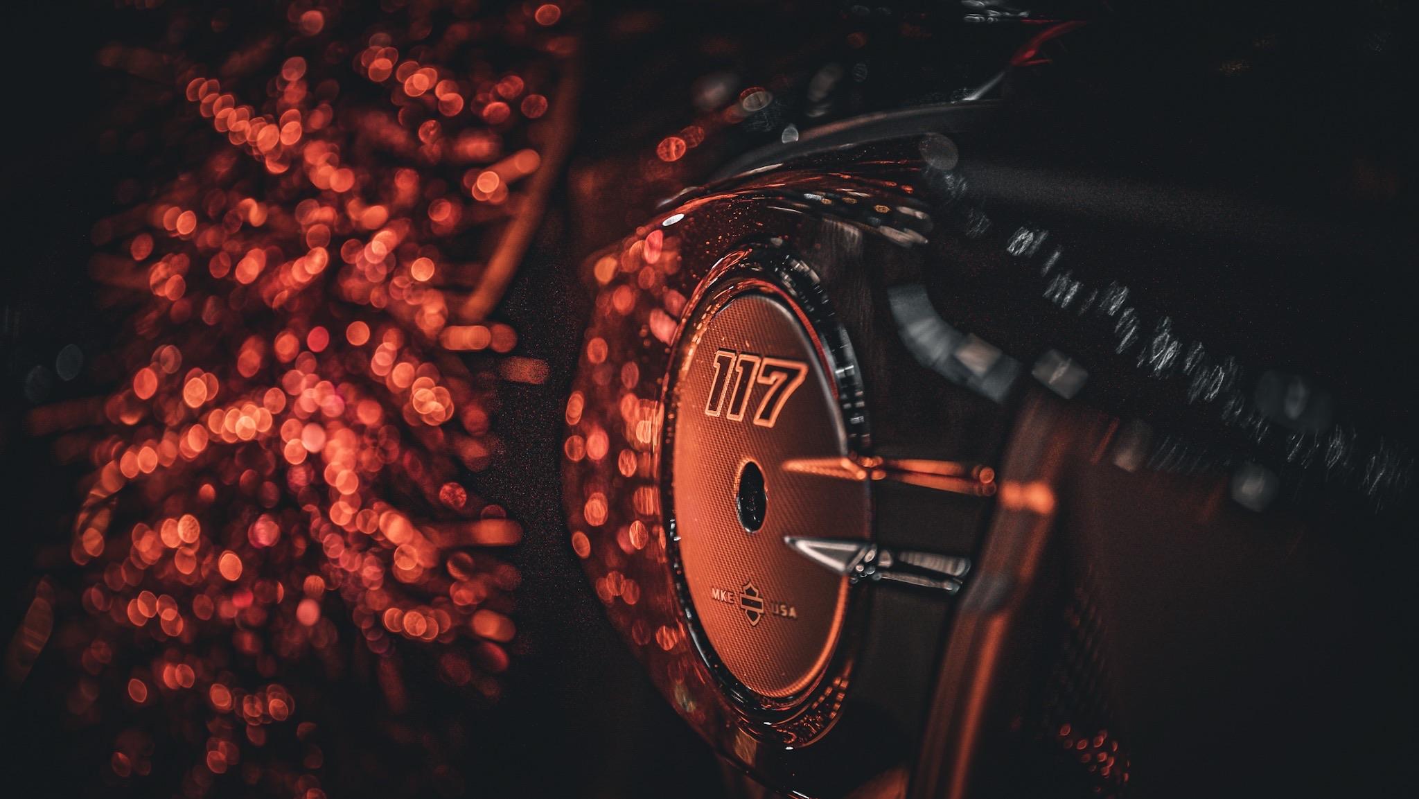 Bliski plan wizerunku emblematu silnika motocyklowego z liczbą „117”, na tle rozmytych czerwonych świateł i efektów bokeh, tworzących dramatyczną i nastrojową atmosferę.