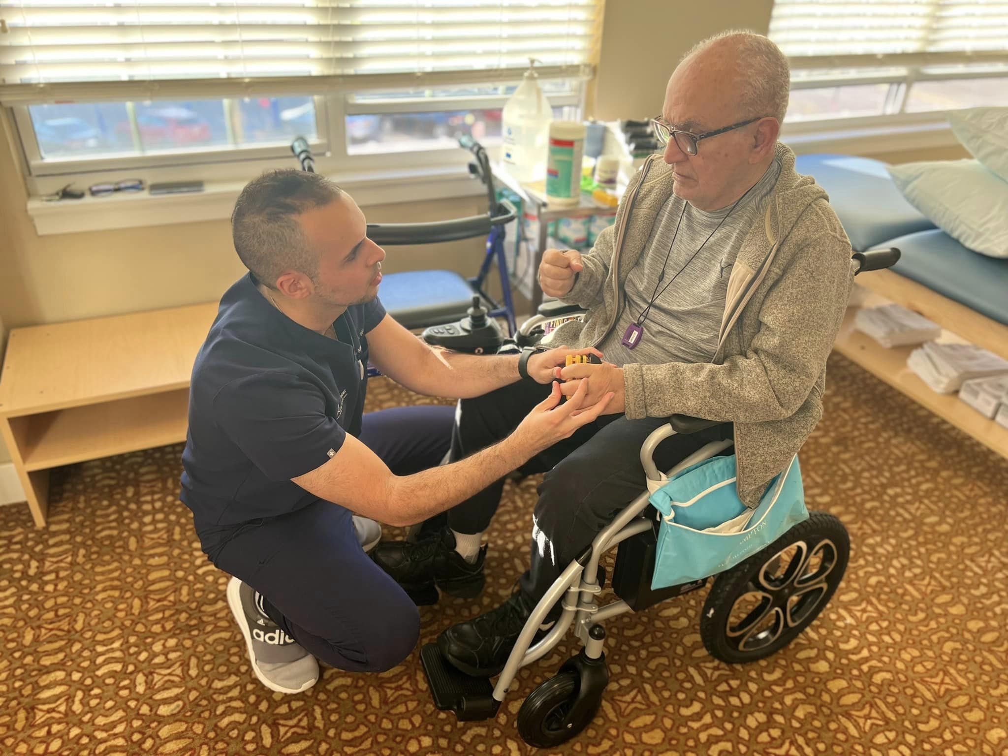 A physical therapist in a subacute rehab facility with his hands on a senior's hands while the senior sits in a wheel chair